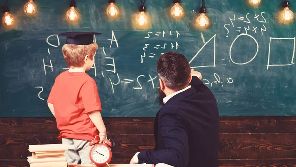 Teacher and schoolboy turned back in front of the green board. Kid in graduation cap learning shapes. Adult pointing at triangle drawn on the board — Stock Photo, Image