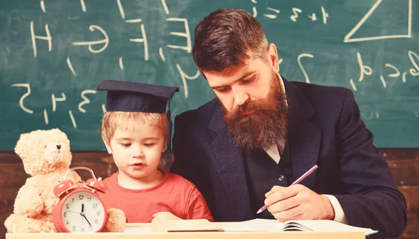 Individual lesson with first grader. Teacher and pupil in mortarboard in classroom, chalkboard on background. Individual studying concept. Teacher, father checking homework, helps to boy, son