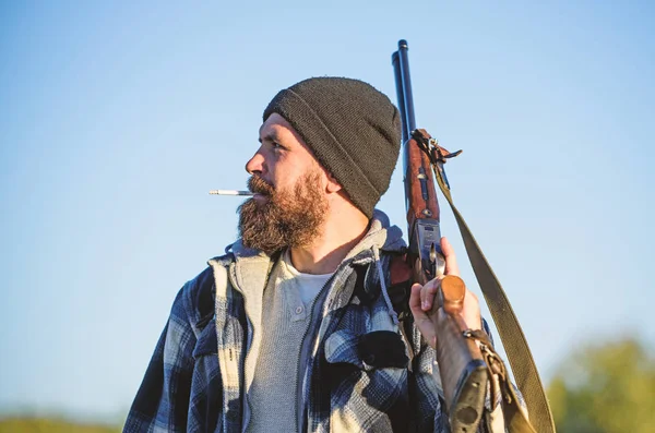 Man brutal bearded guy gamekeeper blue sky background. Hunter with rifle gun close up. Guy bearded hunter spend leisure hunting and smoking. Brutality and masculinity. Hunting masculine hobby concept