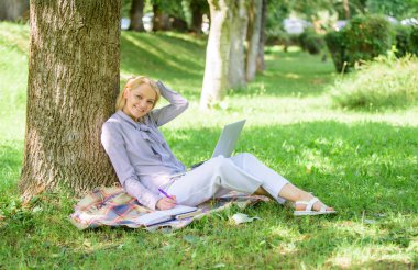 Dizüstü bilgisayarı olan kadın açık havada ağaç gövdesine yaslanır. Eğitim teknolojisi ve internet konsepti. Kız parkta dizüstü bilgisayar ile çim üzerinde oturup çalışır. Doğal çevre ofisi. Açık havada çalışmanın avantajları