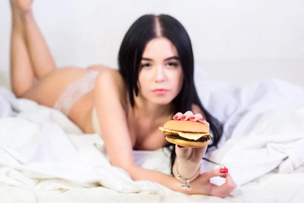 Eating junk food in bed. Girl in sexy lingerie eat burger. Sexual appetite. Food delivery service. Seductive sexy woman relax on bed. Diet concept. Fast food. Seductive woman hungry. Lazy day food