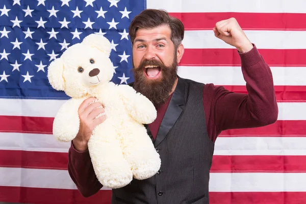 Feliz día de la independencia. Feliz hipster sosteniendo juguete de oso de peluche sobre fondo de bandera americana. Hombre barbudo con cara feliz y emocional celebrando el 4 de julio. Es tan feliz. — Foto de Stock