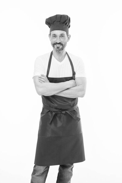 Держать руки скрещенными с уверенностью. Взрослый мужчина в шляпе шеф-повара и фартуке. Старший повар с бородой и усами в фартуке. Пожилой шеф-повар в красном фартуке. Домашняя кухня
