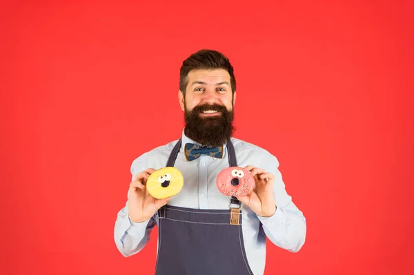 Hipster barbu boulanger tenir des beignets. Doux beignet. Serveur dans un café. Calories de beignets. Un beignet glacé. Homme barbu bien entretenu dans un tablier vendant des beignets. Nourriture aux beignets. Des pâtisseries. Bonbons et gâteaux. La malbouffe — Photo