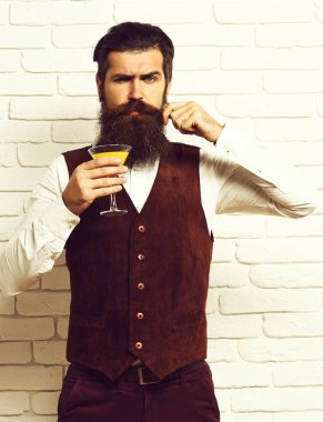 Uzun sakallı ve bıyıklı yakışıklı bir adamın ciddi yüzünde şık saçları var. Beyaz tuğla duvarlı stüdyo arka planında klasik süet deri bir yelek içinde alkollü içecek bardağı tutuyor.