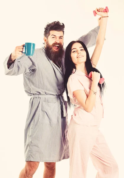 Couple, family on sleepy faces, full of energy. Couple in love in pajama, bathrobe stand isolated on white background. Morning routine concept. Girl with dumbbell, man with coffee cup