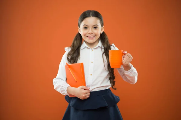 Tout bonheur dépend d'un petit déjeuner scolaire. Petite fille profitant de sa pause scolaire sur fond orange. Mignon écolier souriant avec tasse et carnet de notes. Heureuse écolière ayant une boisson chaude à l'école — Photo