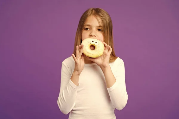 Mädchen mit großen Donut mit Zuckerguss. süße Leckerei, glasierter Bagel mit Augen. Kind isst lustiges Dessert, süße Kindheit. Kind mit langen Haaren trägt weißes Outfit auf violettem Hintergrund — Stockfoto