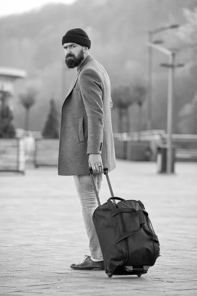 Carry travel bag. Man bearded hipster travel with luggage bag on wheels. Adjust living in new city. Traveler with suitcase arrive airport railway station urban background. Hipster ready enjoy travel