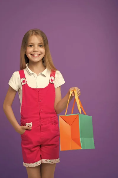 Szczęśliwa dziewczyna z torbą na zakupy na fioletowym tle. Shopaholic mały uśmiech z papierowej torebki. Dziecko shopper w kombinezon mody. Przygotowania do Świąt i uroczystości. Dzieciństwa. Zakupy i sprzedaż — Zdjęcie stockowe