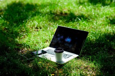 Kahve molası dışarıda. Yaz parkı. Doğal ortamda çalışın ve rahatlayın. Kahve zamanı. Kahve yiyin. Yeşil çim üzerinde kulaklık ve kahve fincanı ile Laptop modern akıllı telefon. Yeşil ofis