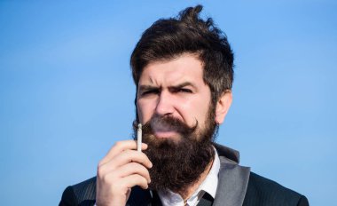 Sakallı ve bıyıklı bir adam sigara tutuyor. Sakallı hipster sigara mavi gökyüzü arka plan sigara. Sigaralı adam nikotin etkisinin tadını çıkarıyor. Sigara içmenin zevki. Rahatlatıcı ama zararlı alışkanlık