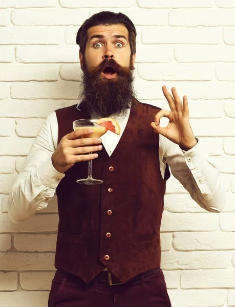 Vousatý muž s dlouhým plnovousem a knírkem má stylové vlasy na překvapené tváři se sklenicí alkoholických nápojů ve starožitném koženém vestě o bílém cihlovém nástěnném studiu backgroun — Stock fotografie