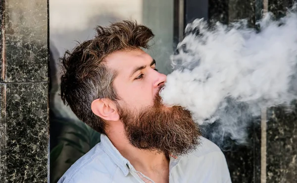 Smoking electronic cigarette. Stress relief concept. Smoking device. Man long beard relaxed with smoking habit. Clouds of flavored smoke. Bearded man smoking vape. Man with beard breathe out smoke