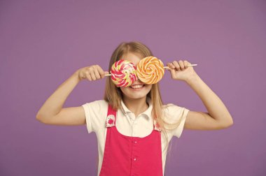 Lolipop menekşe arka plan üzerinde komik çocuk. Şeker gözlerle gülümseyen kız. Küçük çocuk gülümseme şekerler üzerinde sopa ile. Tatlı görünüyorsun. Candyshop kavramı. Çocukluk ve mutluluk. Şeker ile eğleniyor