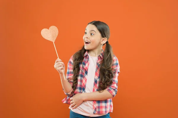 I have my valentines. Adorable valentines girl on orange background. Little child looking at prop heart on valentines day. Cute kid holding fancy valentines day party props on stick — Stock Photo, Image
