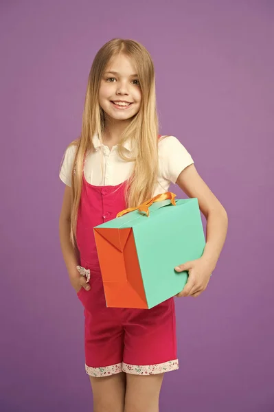 Dziewczyna z uśmiechem dzierżawa worek kolorowy papier. Model z pole pomarańczowy i zielony prezent. Dziecko z długimi blond włosami na sobie kombinezon różowy. Piękne dziewczyny na białym tle na fioletowym tle, koncepcja moda — Zdjęcie stockowe