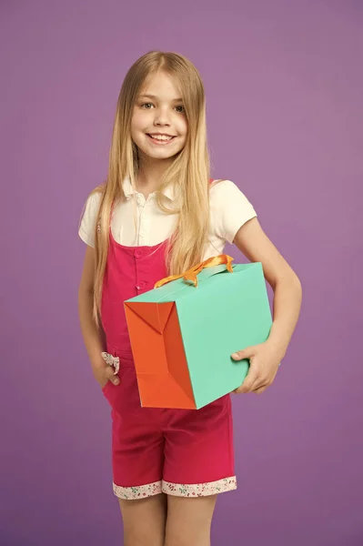 Fille avec un grand sourire tenant sac en papier coloré. Modèle avec boîte cadeau orange et verte. Enfant aux longs cheveux blonds portant une salopette rose. Belle fille isolée sur fond violet, concept de mode — Photo