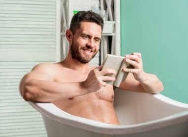 Banyoda seksi adam okuyor. Küvette maço çıplak. Seks ve rahatlama konsepti. adam kaslı bir vücuda sahiptir. Köpükleri suyla dikkatlice yıkayın. kitap sever. banyoda maço zevk kitap. mutlu adam