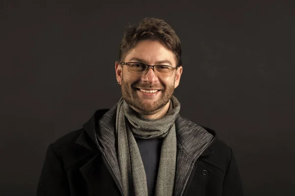 Bearded man in scarf and coat on dark background. Happy man in glasses on bearded face. Fashion model with beard smile on dark background. Autumn style and trend. Fashion accessory concept