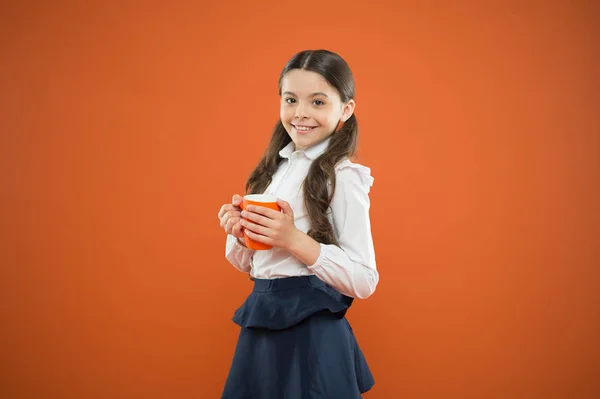 Es hora de descansar. Linda colegiala disfrutando de las vacaciones escolares en fondo naranja. Niña tomando té en la escuela. Pequeño niño sosteniendo la taza de beber durante el descanso — Foto de Stock