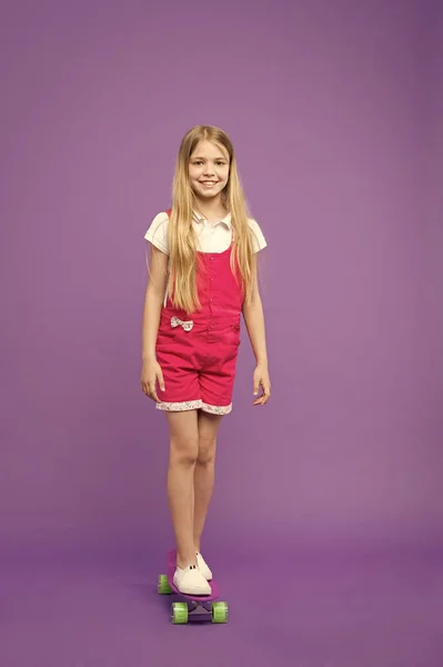 紫色の背景に分離されたスケート ボード上の子。楽しみを持っているピンクのオーバー オールの女の子。スポーティな子供のバランス、アクティブなレジャーの概念。長いブロンドの髪のスリムな女の子のスケート ボードが好き — ストック写真