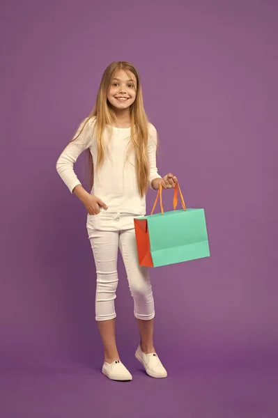 Chica con jersey blanco y pantalones aislados sobre fondo púrpura. Niño con cara alegre sosteniendo bolsa de papel. Niño sorprendido por el regalo, concepto de cumpleaños. Chica con gran sonrisa haciendo compras —  Fotos de Stock