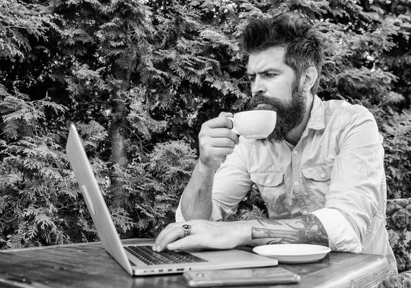 Balancing between work and pleasure. Hipster drinking tea and using computer work station outdoor. Bearded man doing his work online. Working globally through distance work