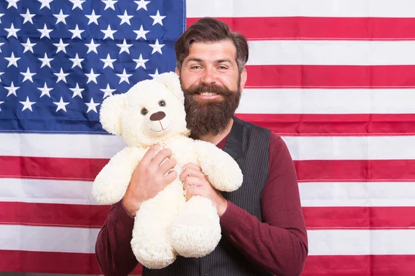 Homem barbudo hipster segurar ursinho de pelúcia fundo bandeira americana. Cuidados infantis. Ainda é infantil. 4 de Julho. Dia da independência. O americano com brinquedo celebra as férias. Férias americanas. Adopção no estrangeiro — Fotografia de Stock