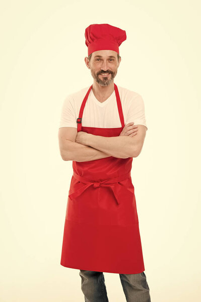 Держать руки скрещенными с уверенностью. Взрослый мужчина в шляпе шеф-повара и фартуке. Старший повар с бородой и усами в фартуке. Пожилой шеф-повар в красном фартуке. Домашняя кухня
