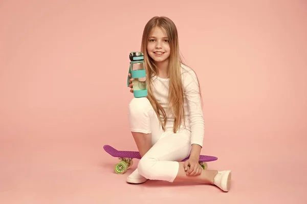 Chica cara feliz sostiene con botella de agua mientras se sientan penny board fondo rosa. Niña cuidando la hidratación corporal. Ocio activo y equilibrio hídrico. Los niños activos y sanos beben agua. Mantenerse hidratado — Foto de Stock