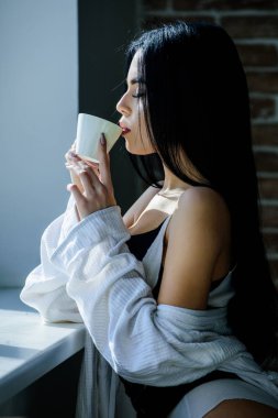 Günlük rutini için en iyi kahve. Şehvetli kız en sevdiği sabah kahvesini içiyor. Güzel bir kadın pencerede taze sıcak kahve içiyor. Seramik kahve fincanı tutan seksi kız