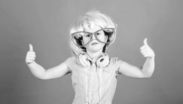 快適なヘッドフォンのための親指アップ。ヘッドフォンとピンクの髪のかつらを着て派手なメガネの小さな女の子。近代的なワイヤレスヘッドフォンを備えた小さな子供。かわいい子供は耳のステレオヘッドフォンを承認 — ストック写真