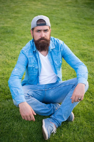 Obejmując estetykę sklep streetwear. Sexy mężczyzna siedzi na zielonej trawie. Brodaty człowieka w stylu casual denim. Kaukaski mężczyzna z wąsy i zarost. Brutalny mężczyzna z nieogoloną twarzą w kapelusza SnapBack — Zdjęcie stockowe