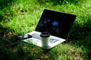 Laptop, kulaklıklı ve yeşil çimenli kahve fincanlı modern akıllı telefon. Kahve molası. Yaz parkı. Doğal ortamda çalış ve rahatla. Yeşil ofis. Kahve zamanı. Kahveni götür.