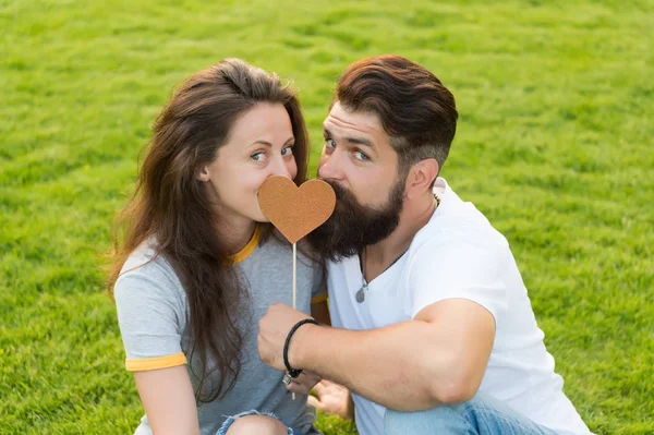 Given his heart to her. Sensual couple in love with prop heart on green grass. Sexy woman and bearded man holding valentines heart on stick. One heart two souls