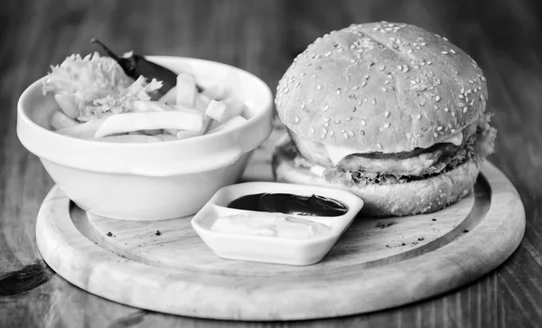 Hamburguesa con carne de queso y ensalada. Comida de engaño. Deliciosa hamburguesa con semillas de sésamo. Menú de hamburguesas. Snack alto en calorías. Hamburguesa y papas fritas y salsa de tomate sobre tabla de madera. Concepto de comida rápida —  Fotos de Stock