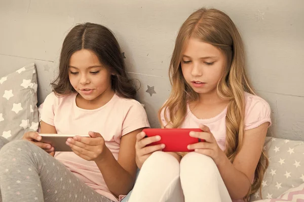 Children in pajama interact with smartphones. Application for kids fun. Internet surfing and absence parental advisory. Smartphone internet access. Girls sisters wear pajama busy with smartphones