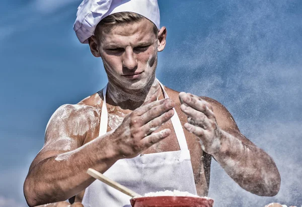Chef cook preparing dough for baking with flour. Man on busy face wears cooking hat and apron, sky on background. Cook or chef with sexy muscular shoulders and chest covered with flour. Baker concept