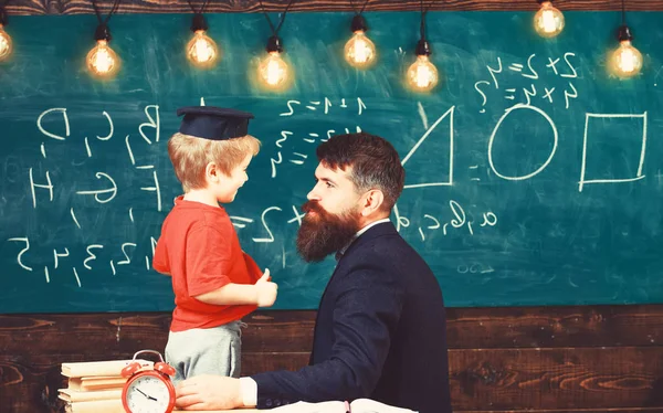 Teacher with beard, father teaches little son in classroom, chalkboard on background. School break concept. Boy, child in graduate cap play with dad, having fun and relaxing during school break