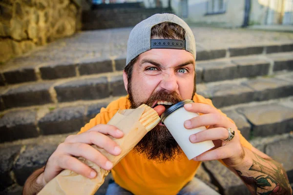 Snack a jó hangulat. Guy evés hot dog. Utcai élelmiszer-koncepció. Ember szakállas enni ízletes kolbászt és inni papírpohár. A városi életmód táplálkozás. Szemét élelmiszer. Gondtalan csípő enni gyorsétel, míg a lépcsőn — Stock Fotó