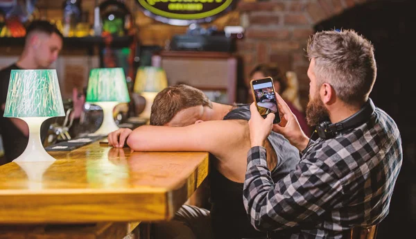 Hipster burlándose de un amigo borracho. Hombre borracho duerme y chico con smartphone. Hipster tomando fotos borracho amigo. Amigos borrachos en el bar. Duerme en el mostrador del bar. Tome una foto para recordar la fiesta —  Fotos de Stock