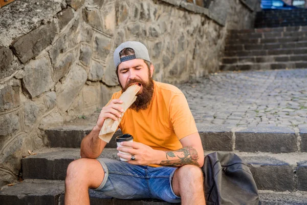 Nutrição de estilo de vida urbano. Despreocupado hipster comer junk food while sit on stairs. Um lanche de homem faminto. Comida de plástico. Um tipo a comer cachorros quentes. Homem barbudo mordida saborosa salsicha e beber copo de papel. Comida de rua tão boa — Fotografia de Stock