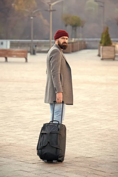 Hipster ready enjoy travel. Carry travel bag. Man bearded hipster travel with luggage bag on wheels. Adjust living in new city. Traveler with suitcase arrive airport railway station urban background