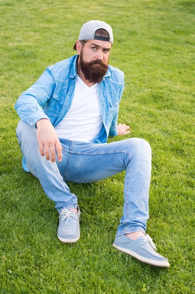 Élégant et confortable à porter. Élégant hipster en casquette de baseball assis sur l'herbe verte en plein air. Homme barbu portant streetwear d'été élégant. Modèle de mode avec look décontracté élégant — Photo