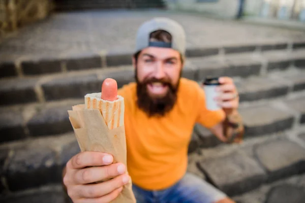 Despreocupado hipster comer junk food while sit on stairs. Um lanche de homem faminto. Comida de plástico. Um tipo a comer cachorros quentes. Homem barbudo mordida saborosa salsicha e beber copo de papel. Comida de rua tão boa. Estilo de vida urbano nutrição — Fotografia de Stock