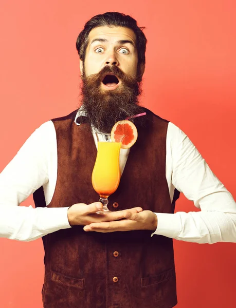 handsome bearded man with long beard and mustache has stylish hair on surprised face holding glass of alcoholic cocktail in vintage suede leather waistcoat on red studio background