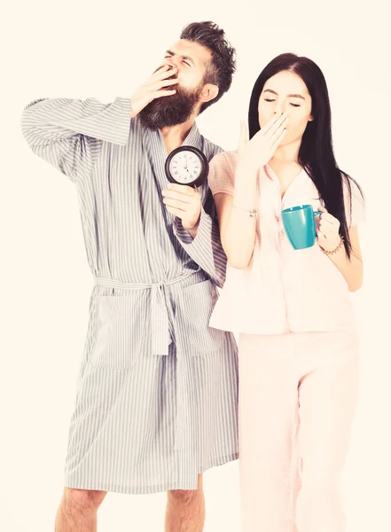 Влюблённая пара, молодая семья в пижаме, халаты изолированы на белом фоне. Девушка с кофейной чашкой, мужчина держит часы в руке и зевает. Идеальная идея для утра. Пара проснулась рано утром — стоковое фото