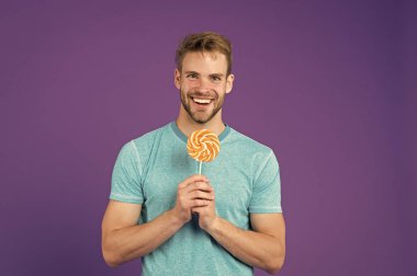 Çocukluk tadı. Tüylü adam lolipopu seviyor. Yemek konseptini aldatmak. Şeker sağlığa zararlıdır. Adamın elinde lolipop şeker menekşesi var. Yakışıklı maço adam kocaman renkli lolipop yiyor.