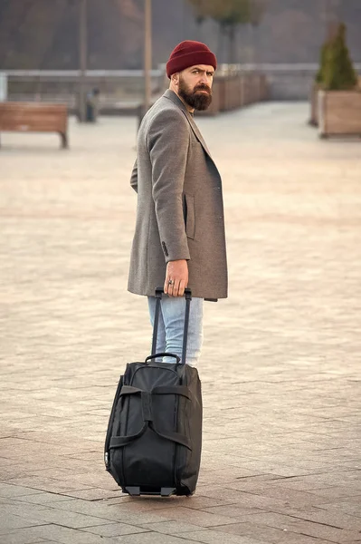 Traveler with suitcase arrive airport railway station urban background. Hipster ready enjoy travel. Carry travel bag. Man bearded hipster travel with luggage bag on wheels. Adjust living in new city